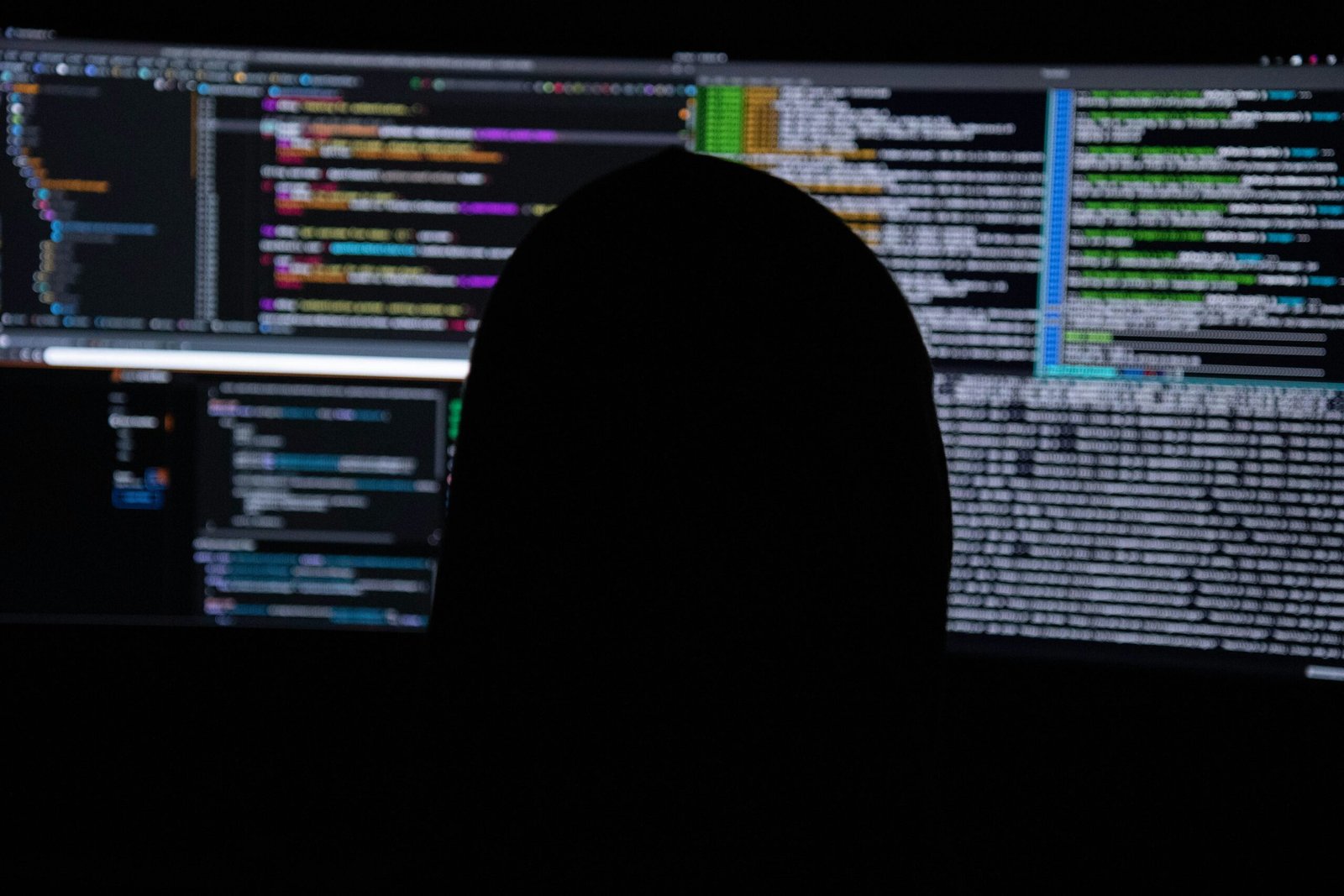 A hooded figure silhouetted against multiple code-filled monitors, symbolizing covert cyber intrusion, command-and-control operations, and ransomware activity.
